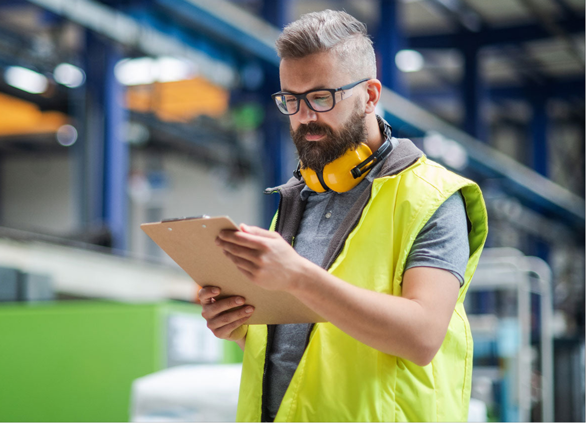 Man in a warehouse wearing a yellow safety vest and glasses, holding a tablet.